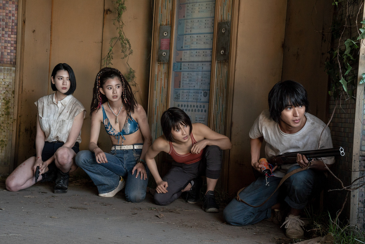  Four young people in a tense moment inside a rundown building, armed and alert, with vines creeping in from the walls, dressed in casual and rugged outfits, prepared for action.