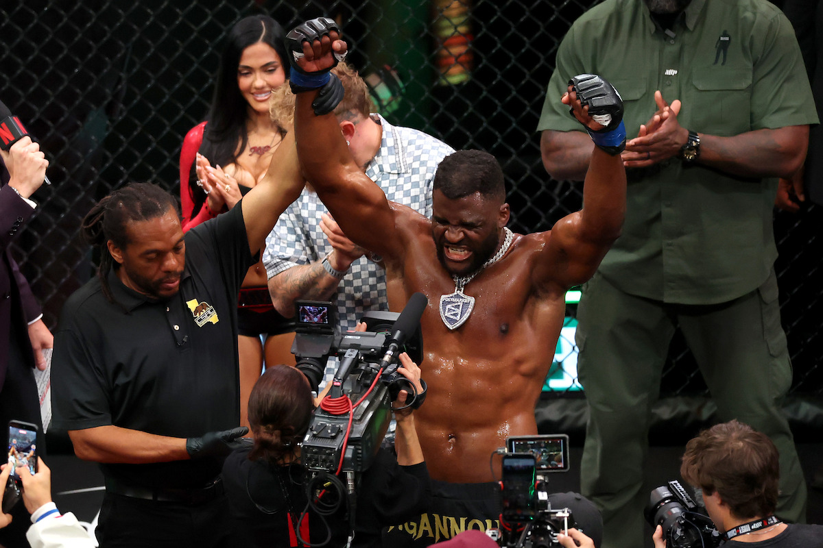 MMA fighter celebrates victory with arms raised inside octagon, surrounded by referee, camera crew, and applauding people. The setting is a professional mixed martial arts event.