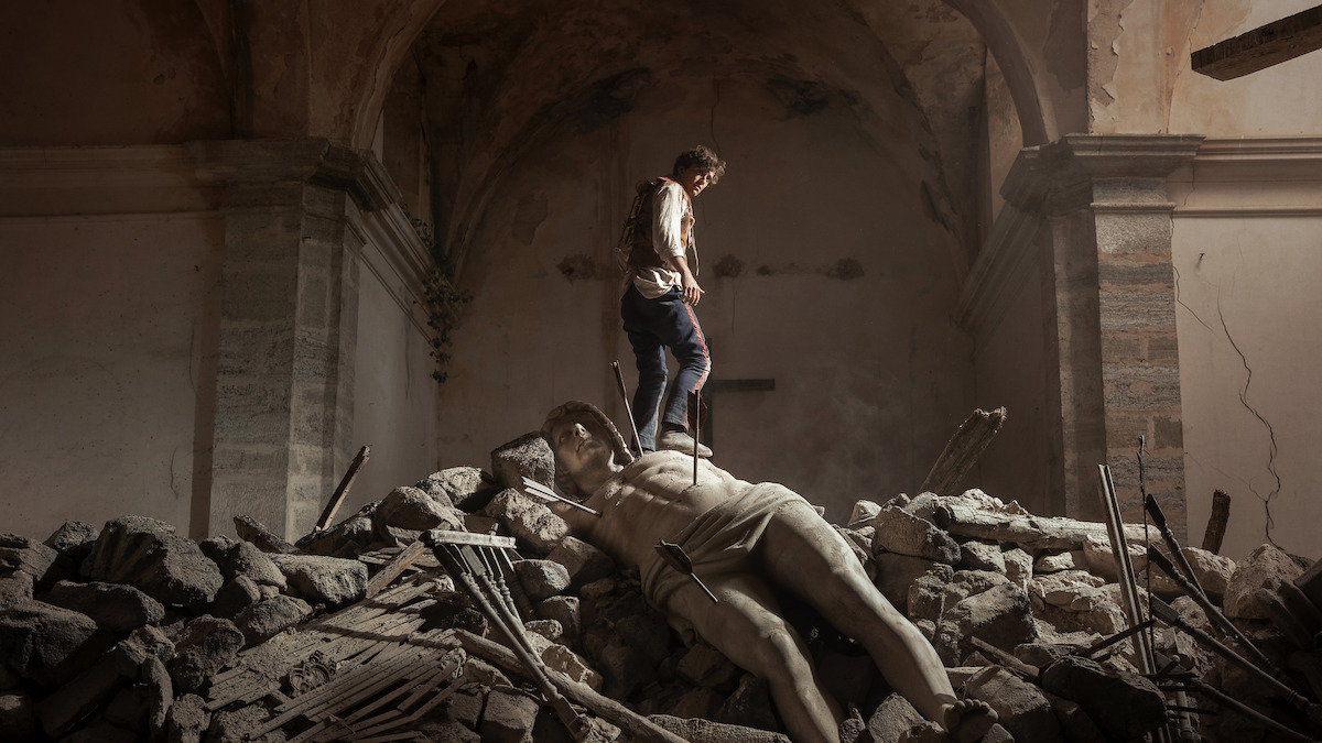 A person standing on rubble inside a damaged, old stone building, with a large fallen classical statue lying among the debris under an arched ceiling, evoking a sense of destruction and loss.