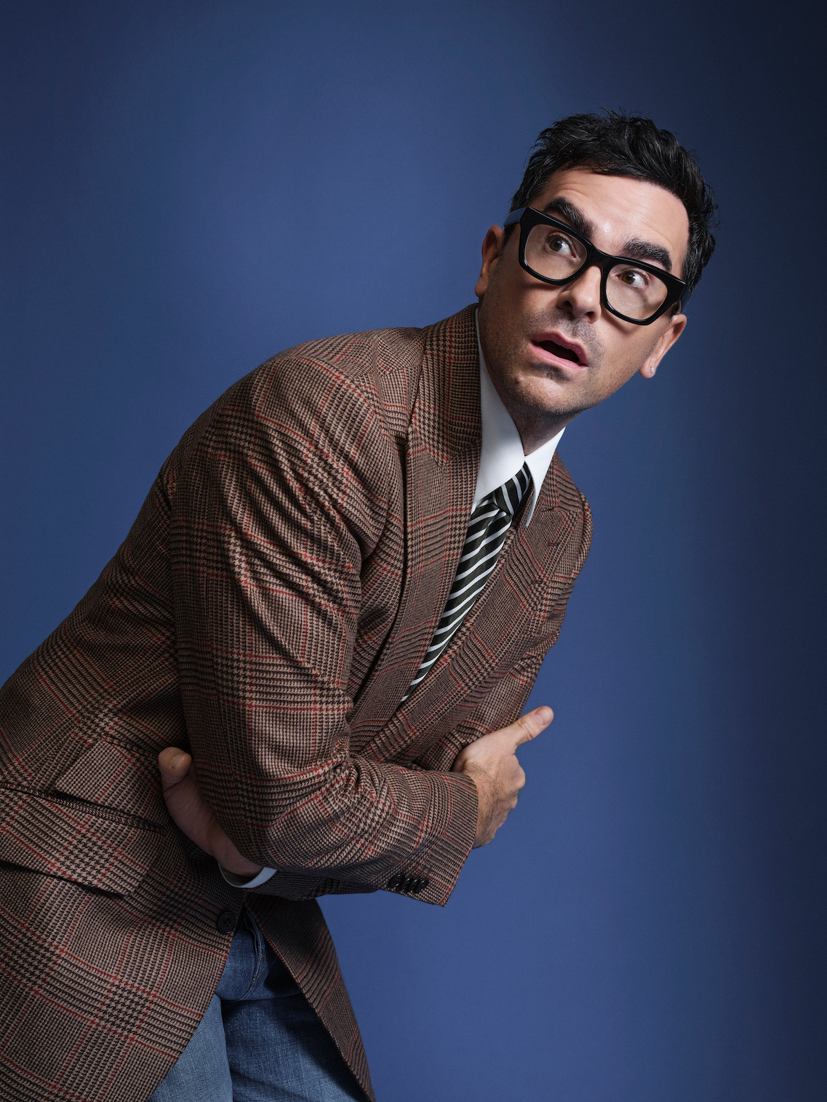 Dan Levy poses against a blue backdrop, wearing a checked blazer.