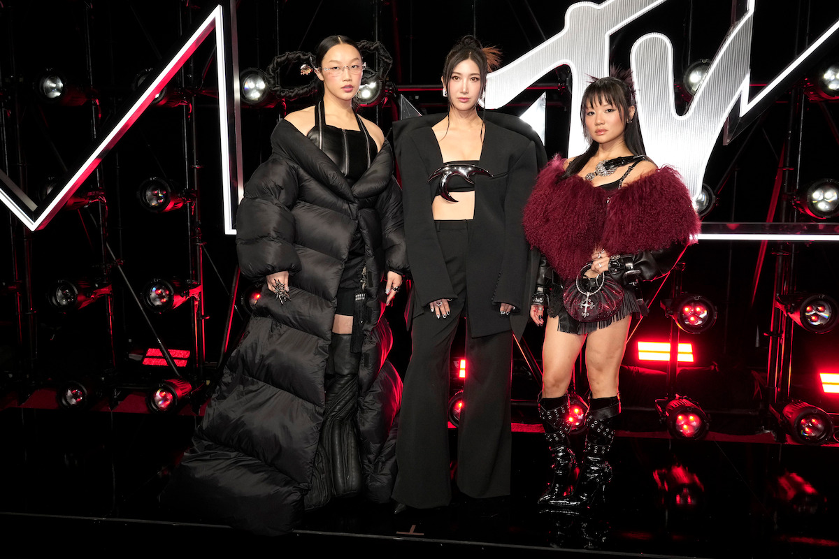 Three women in bold, stylish black and red outfits pose on a red carpet with dramatic lighting and an illuminated MTV logo background at an event.