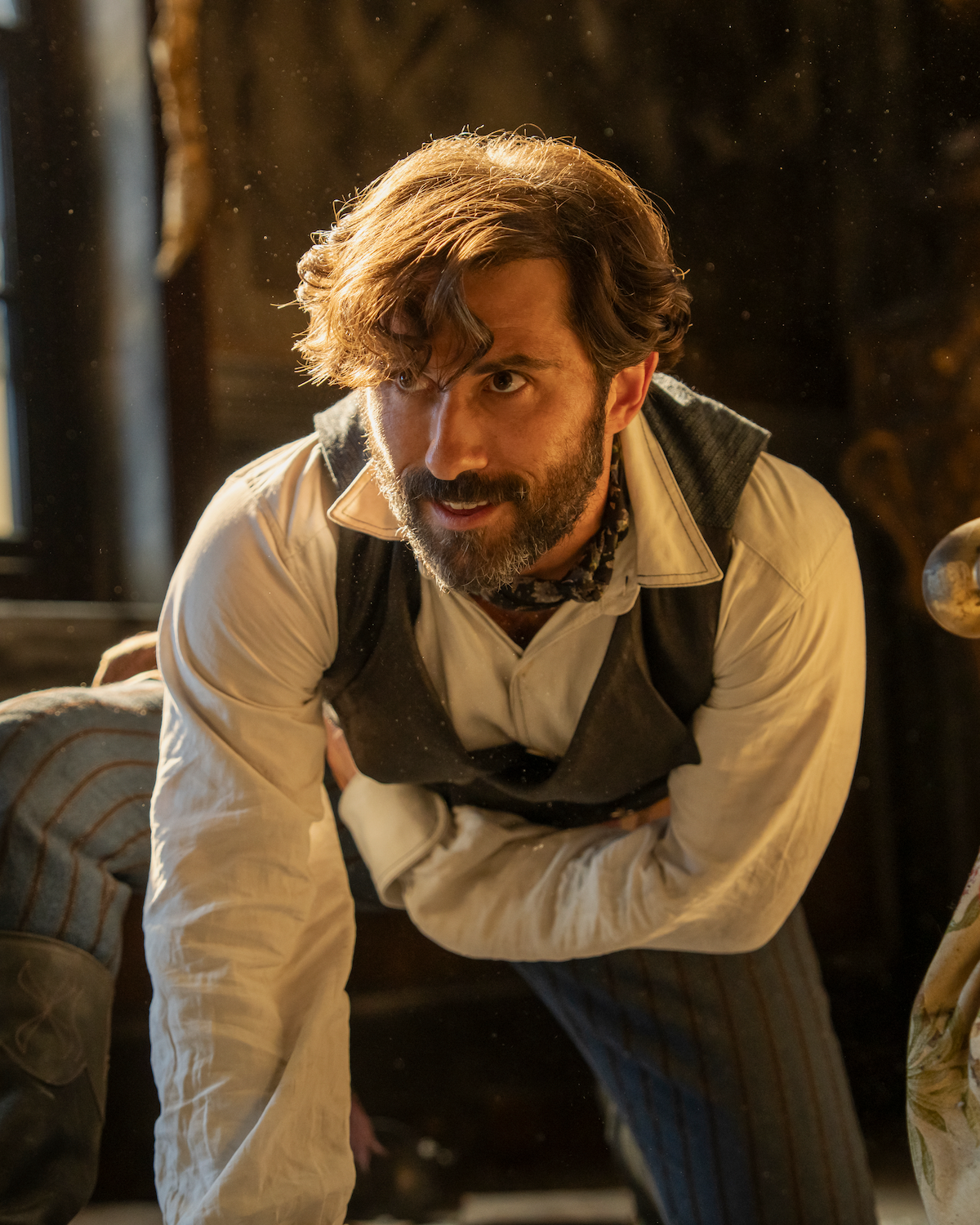 A man with wavy hair and a beard dressed in old-fashioned clothing leans forward in a warmly lit, rustic room, sunlight streaming in from a window behind, suggesting a historical or period drama setting.