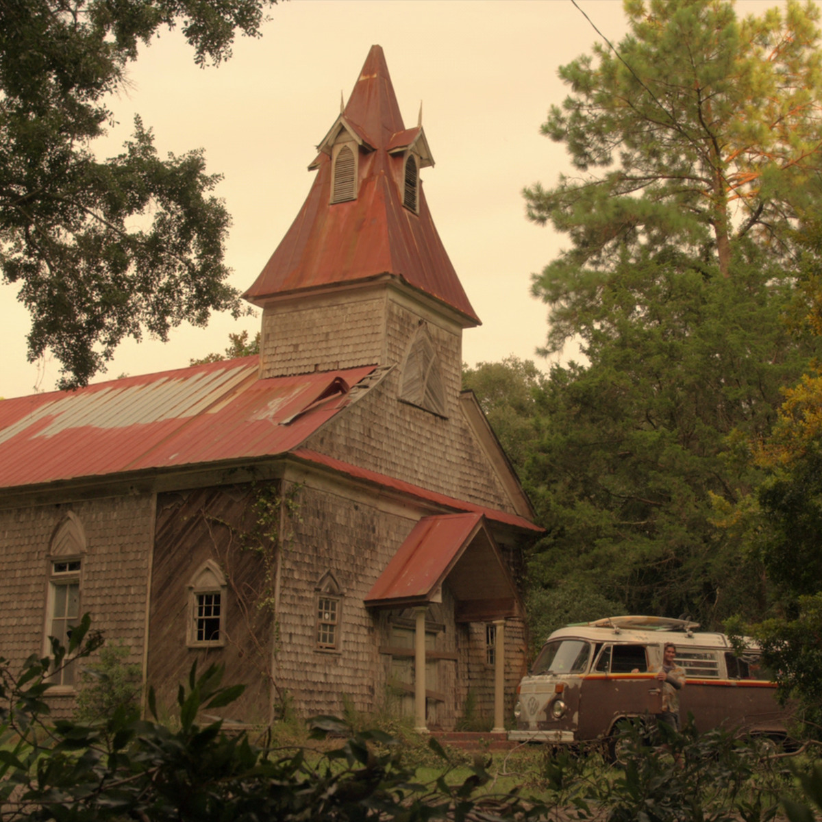 Igreja antiga de madeira com teto vermelho cercada de árvores. Um carro antigo está estacionado ao lado dela sob um céu nublado em um ambiente rural.