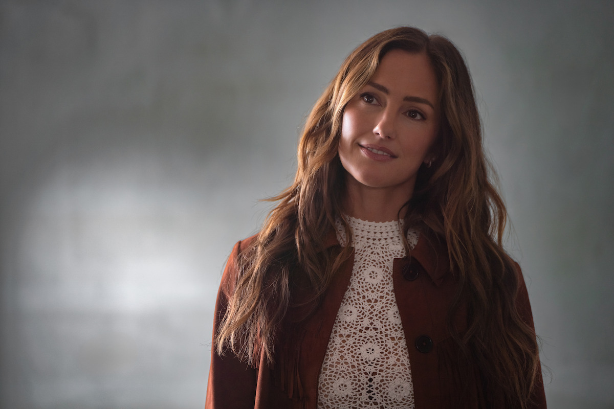Woman with long brown hair wearing a brown jacket and white lace top, standing indoors in front of a blurred gray wall, softly lit, looking slightly to the side with a gentle expression.