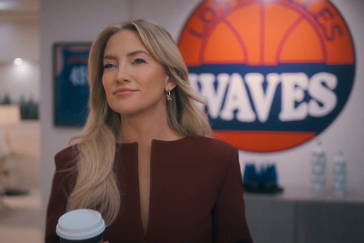 Confident woman with long blonde hair holding a coffee cup in a modern, well-lit office setting with stylish decor and a framed jersey on the wall in the background.