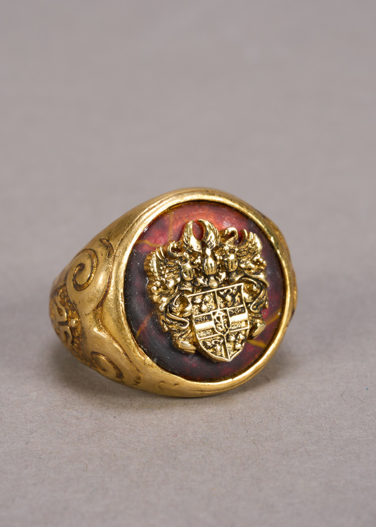 A gold signet ring with engraved crest sits on a plain gray surface under soft lighting, creating an antique, elegant, and historical atmosphere.