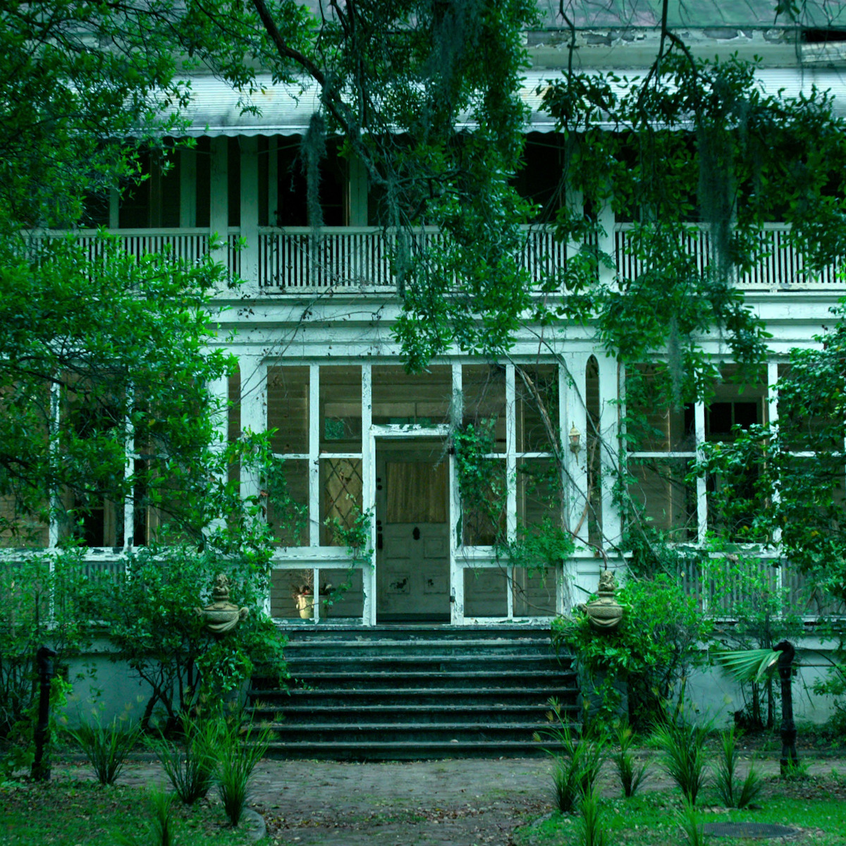 Casa branca antiga com plantas malcuidadas, cercada por uma paisagem verde. O imóvel tem uma varanda na frente, com degraus, e parece abandonado. O ambiente é cercado de vegetação, dando um clima misterioso e assustador.