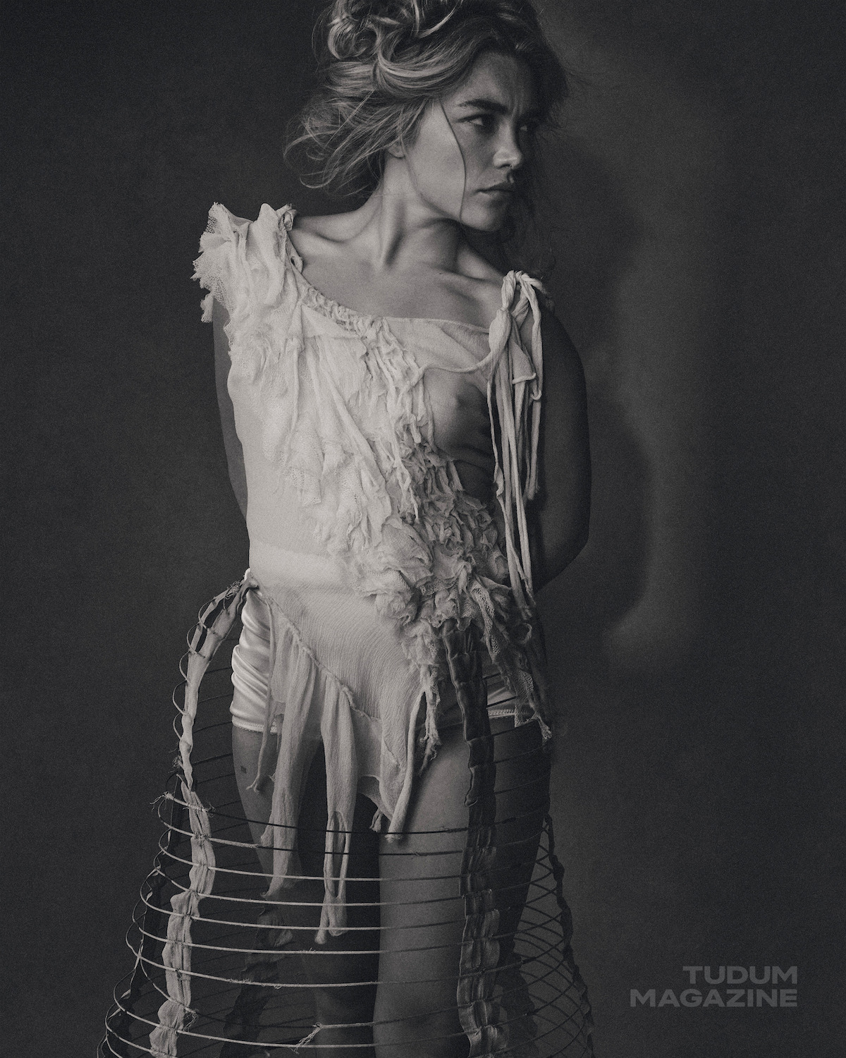 Black and white photo of a woman with styled hair, wearing a distressed, artistic dress with wire skirt elements, standing against a plain dark background, lit with dramatic shadows. "TUDUM MAGAZINE" logo appears in the corner.