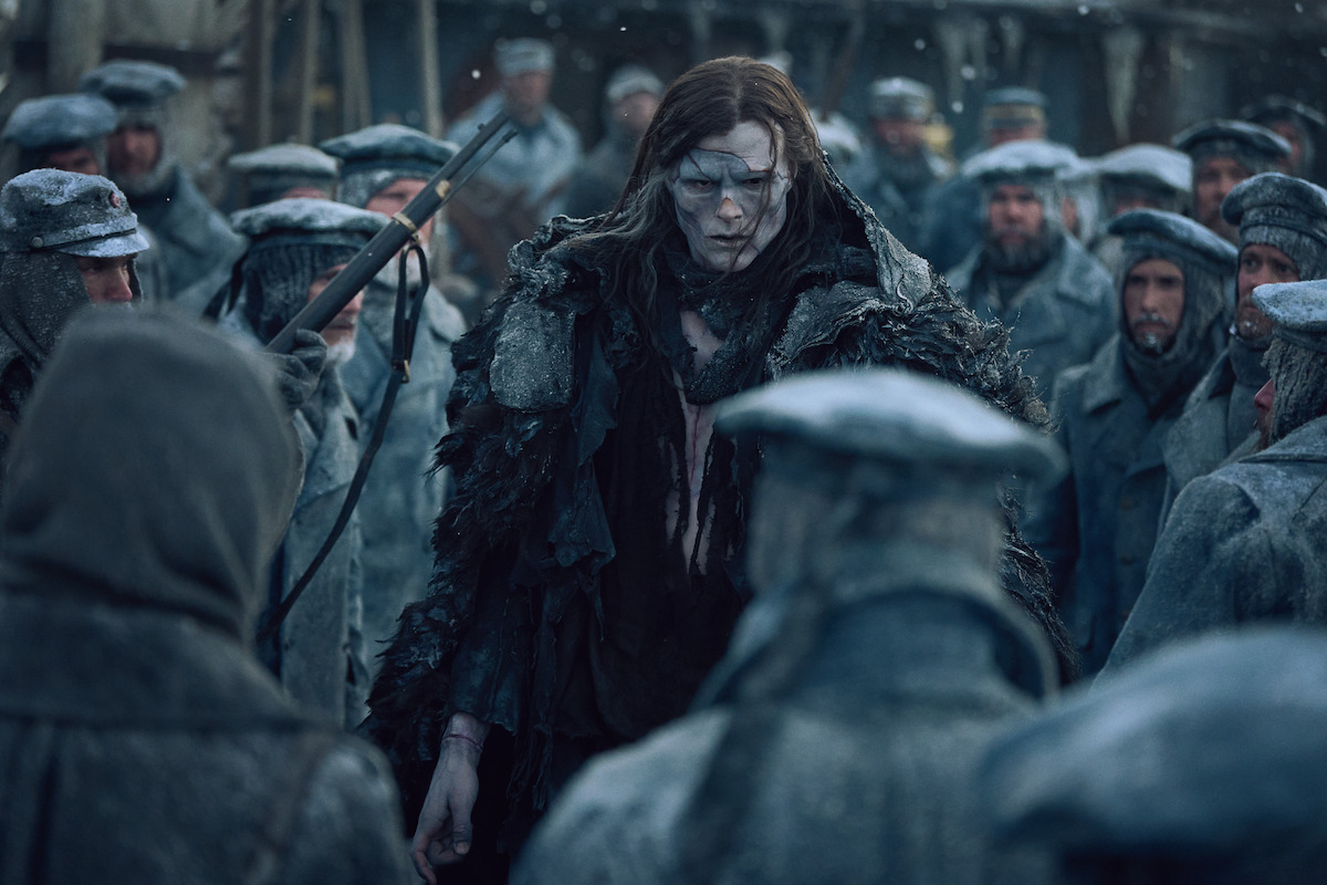 A pale, somber figure in tattered fur stands among a group of uniformed, frost-covered people in a snowy, bleak outdoor setting, evoking a tense and mysterious atmosphere.