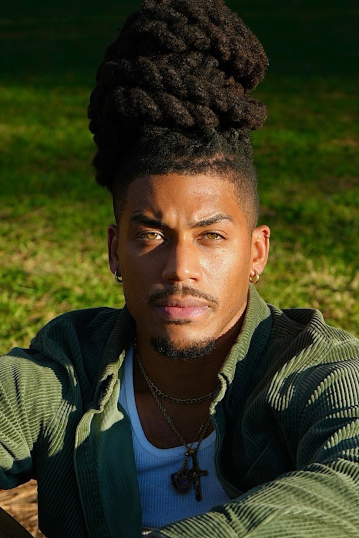 Man with tall dreadlocks and layered necklaces wearing a green jacket and white shirt sits outdoors on grass in sunlight, looking directly at the camera.