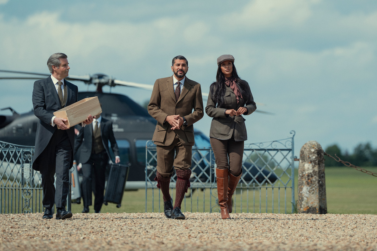 Three people in formal country attire walk on gravel near a helicopter and iron gate on a rural estate, with cloudy sky and grassy field in the background. One carries a box, another has a briefcase.