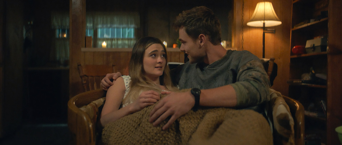 A young couple sits close together on a chair, wrapped in a blanket in a cozy, warmly lit wooden room with a lamp, shelves, and candles in the background, creating an intimate and comfortable atmosphere.