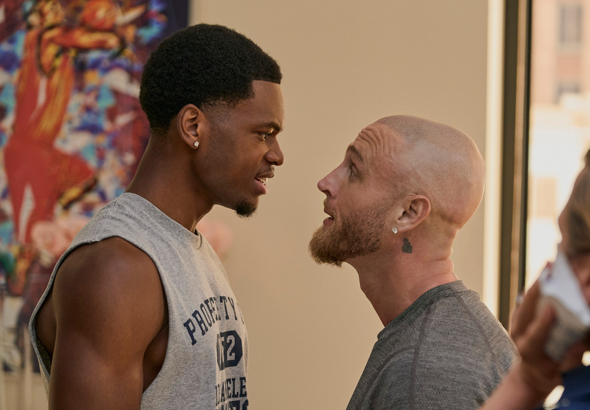 Two men indoors, one holds a framed puzzle of the Golden Gate Bridge at sunset while the other, wearing glasses and a blue jacket, holds up a puzzle piece, indicating it’s the last piece needed to finish the puzzle.