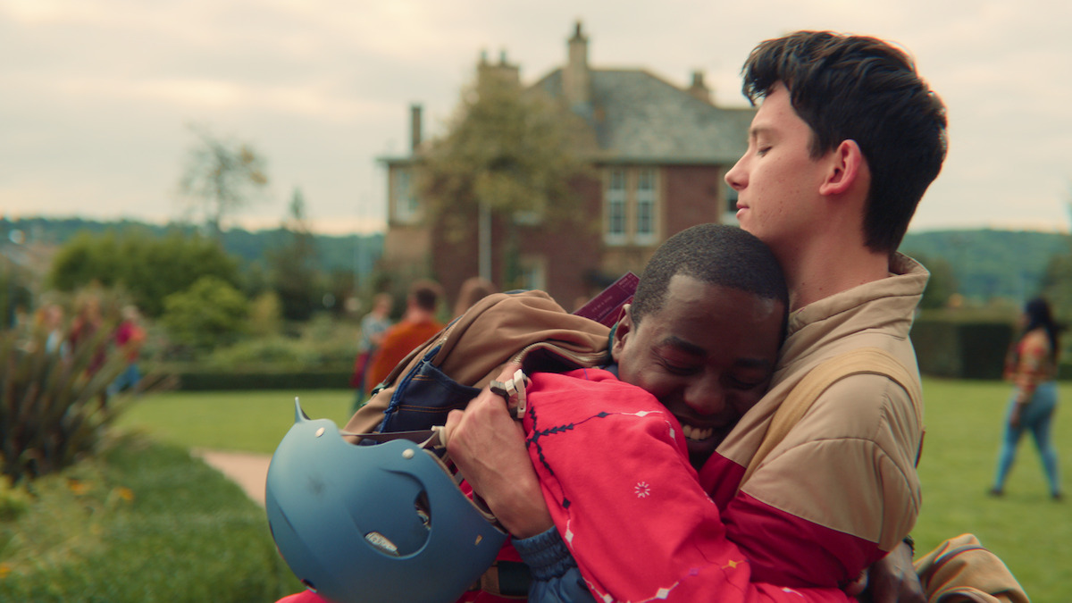 Two friends embrace warmly outdoors with a large house in the background, one smiling and wearing a red jacket, the other with eyes closed, as people socialize on the lawn.