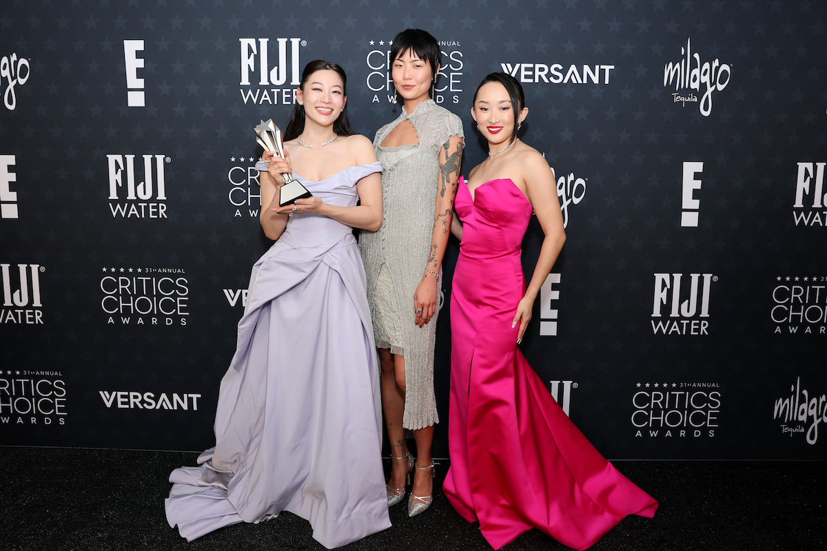 Three women pose on the Critics Choice Awards red carpet, one holding a trophy. They wear elegant gowns in lavender, silver, and bright pink, standing before a backdrop with logos like FIJI Water and Milagro Tequila.