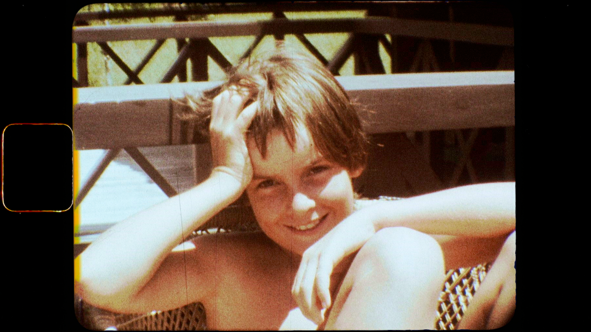 Young person with short hair sitting outdoors on a sunny day, smiling and resting their head on one hand, with wooden fence and greenery in the background, warm and relaxed atmosphere.
