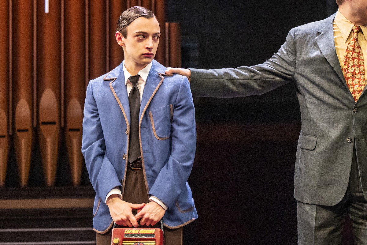 A young man in a blue suit holding a book stands on stage next to an older man in a gray suit with his hand on the young man's shoulder, with organ pipes in the background. The scene appears to be from a theatrical performance.