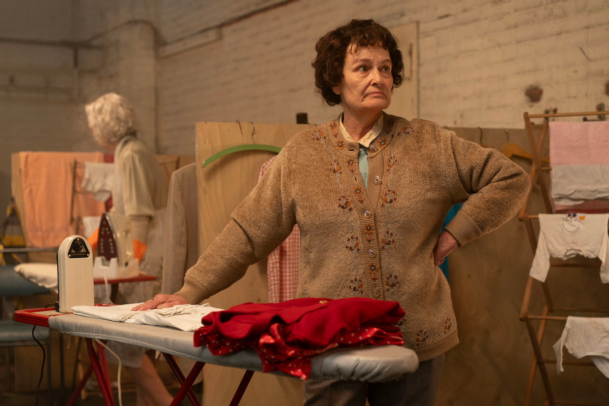 Middle-aged woman standing at an ironing board with red clothing, in a laundry room with drying racks, ironing supplies, and industrial-style brick walls. She has a thoughtful expression and wears a brown sweater.