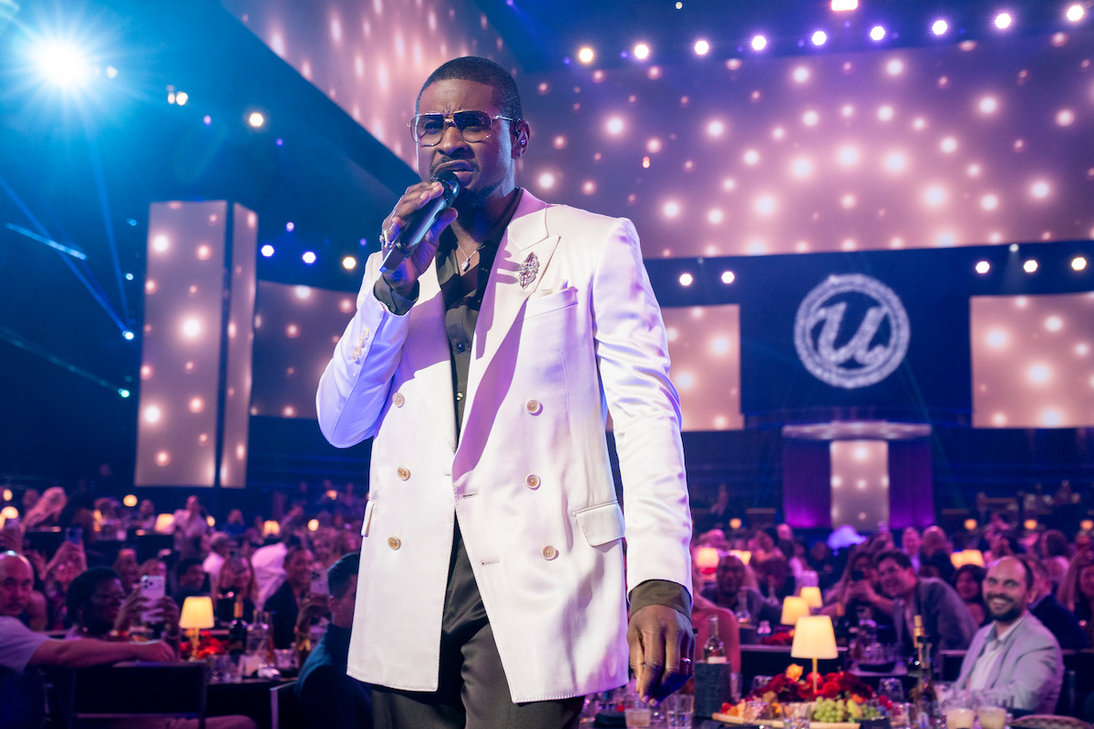 A male singer in a white jacket performs with a microphone on stage at a formal event, surrounded by an audience seated at tables with decorative lights and a large illuminated backdrop.