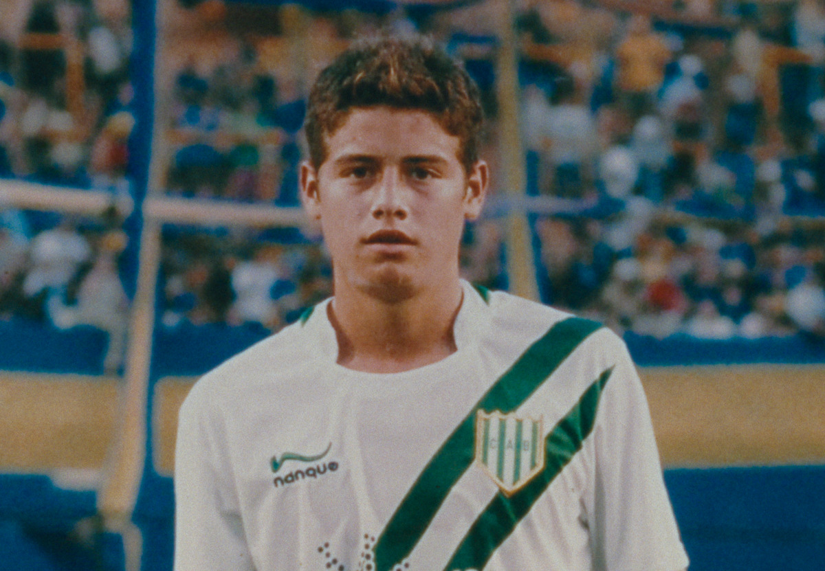 Young male soccer player in a white and green striped jersey stands on a football field, stadium with blue and yellow seats filled with spectators in the blurred background.
