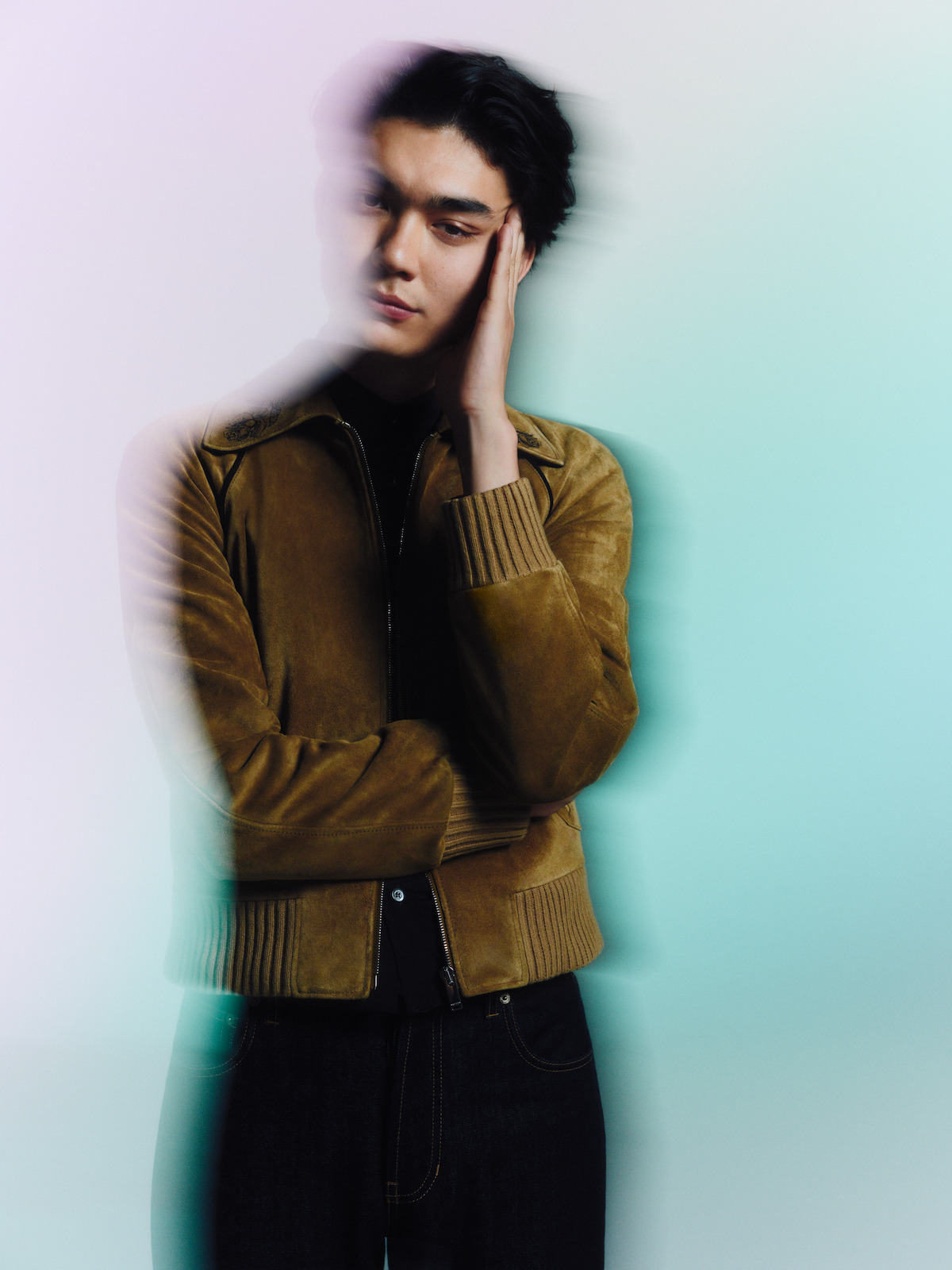 A person in a brown jacket poses against a softly colored, blurred background. The head and hand appear motion-blurred, creating an artistic, dreamy effect in the studio setting.