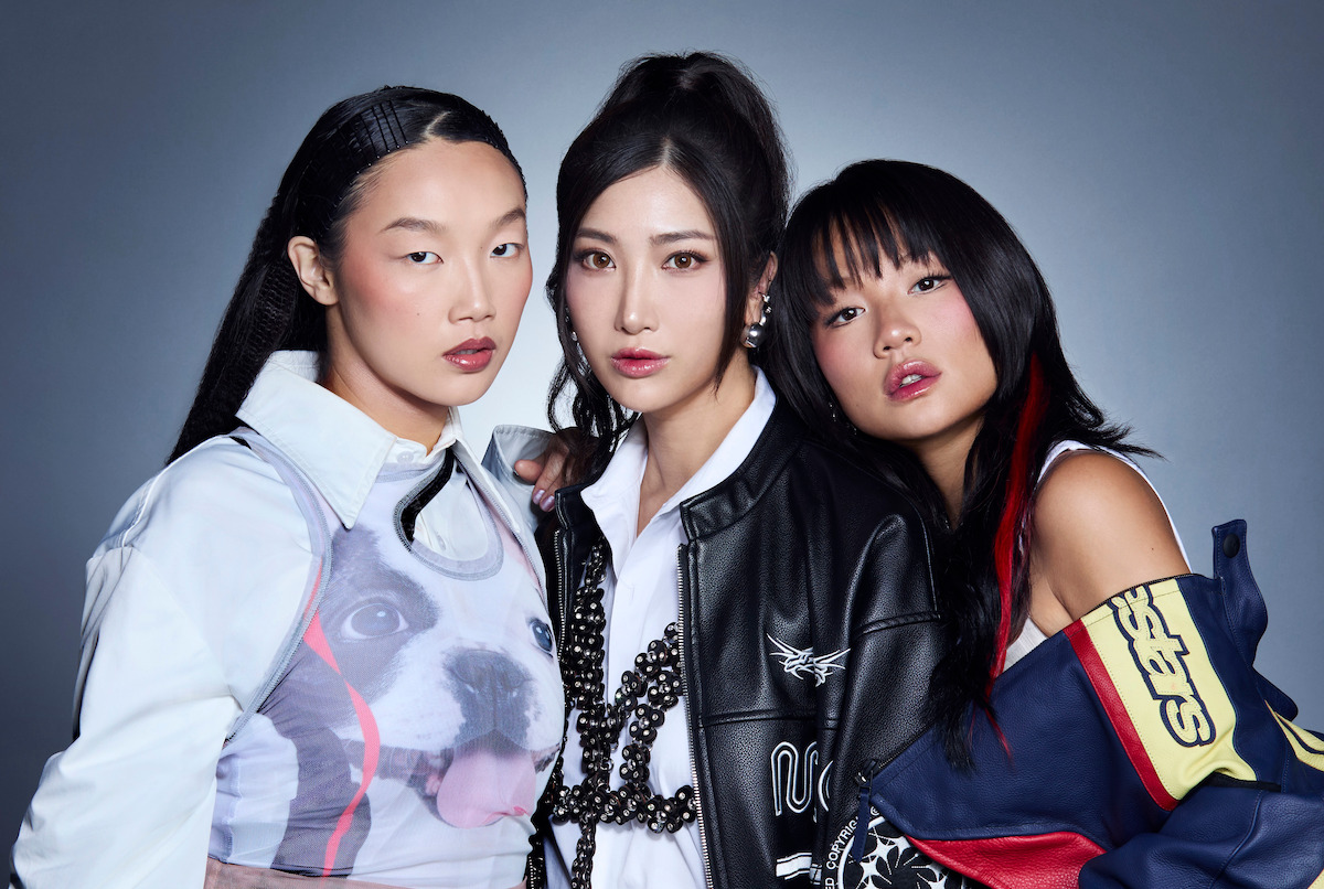 Three fashionable women posing closely together against a plain gray background, wearing trendy, modern outfits with expressive makeup and diverse hairstyles, creating a stylish, confident group portrait.