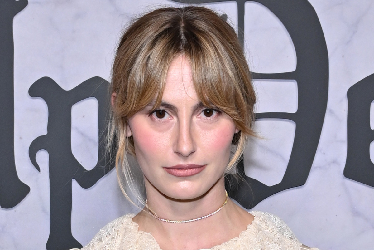 Woman with blond hair and bangs poses indoors against a marble wall backdrop with large black letters. She wears a lace top and a thin necklace, looking directly at the camera with a neutral, composed expression.
