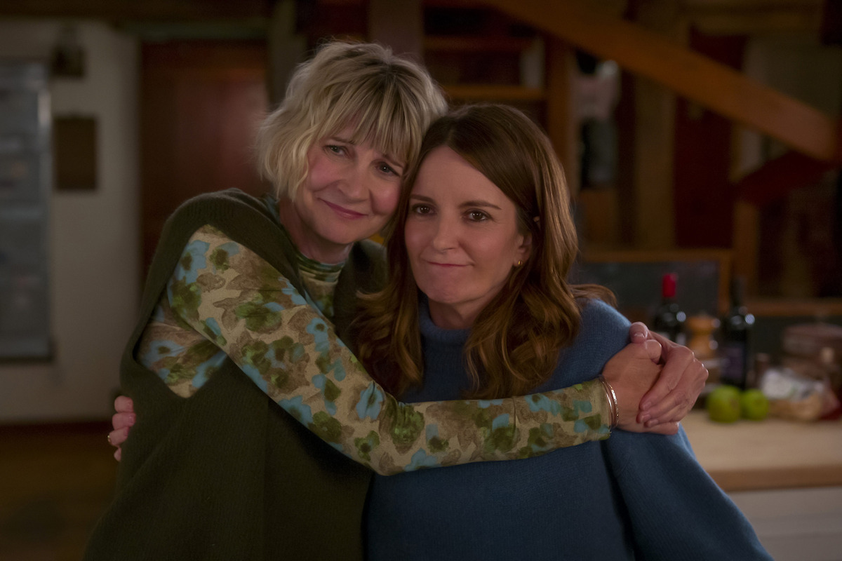 Two women smiling and hugging each other in a cozy, warmly lit kitchen setting with fruit and wine on the counter, conveying a sense of friendship and comfort.