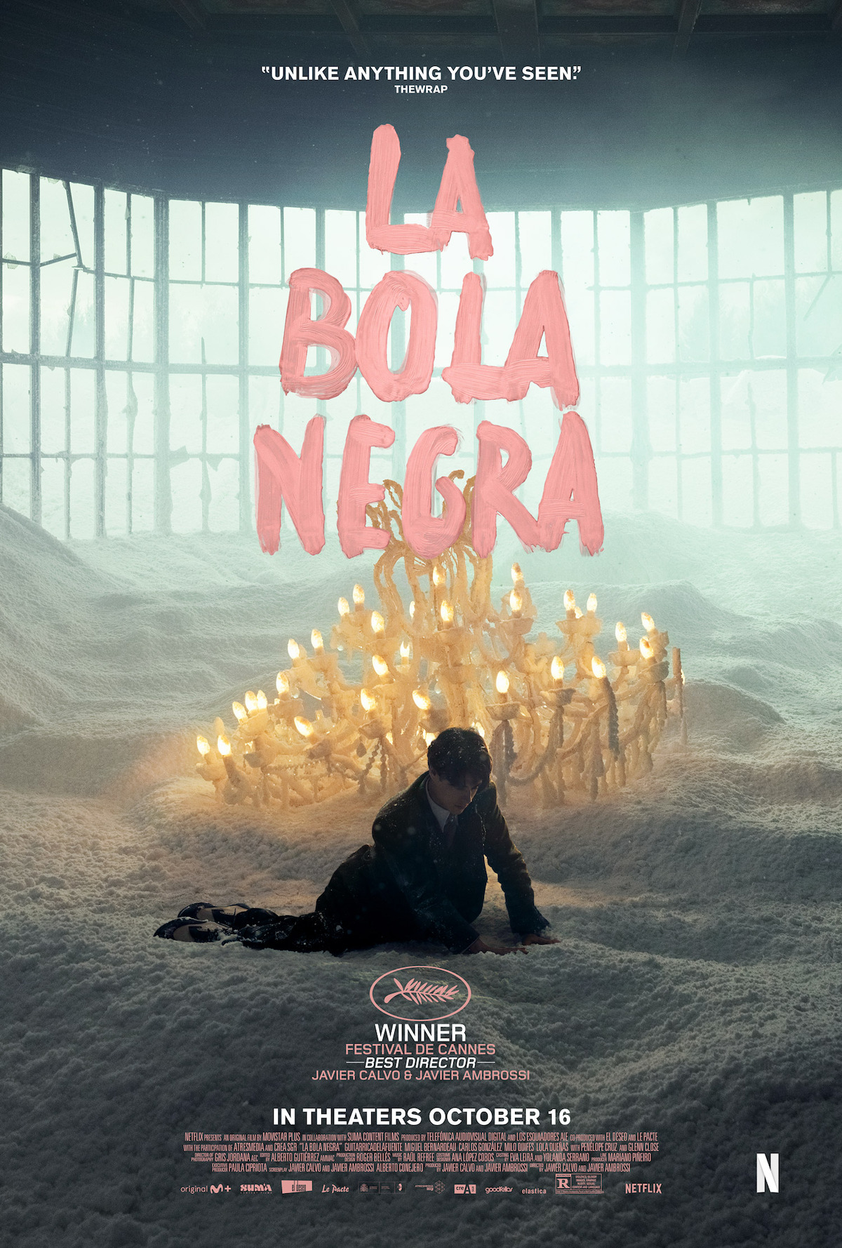 A person kneels in artificial snow beside a fallen chandelier inside a large, windowed room. The setting is surreal, with soft lighting and large pink text reading "La Bola Negra" above. Film festival awards and Netflix logo are shown.