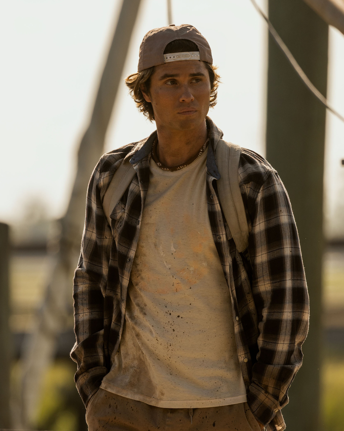 Chase Stokes as John B Routledge, wearing a backward cap, plaid shirt, and backpack stands outdoors in a sunlit, rustic environment with wooden posts and blurred background, looking off into the distance.
