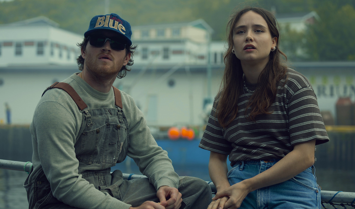 Two people sit outdoors near a harbor, with fishing docks and buildings in the background. The man wears sunglasses, overalls, and a cap, while the woman wears a striped shirt and jeans. Both appear to be looking at something off-camera.