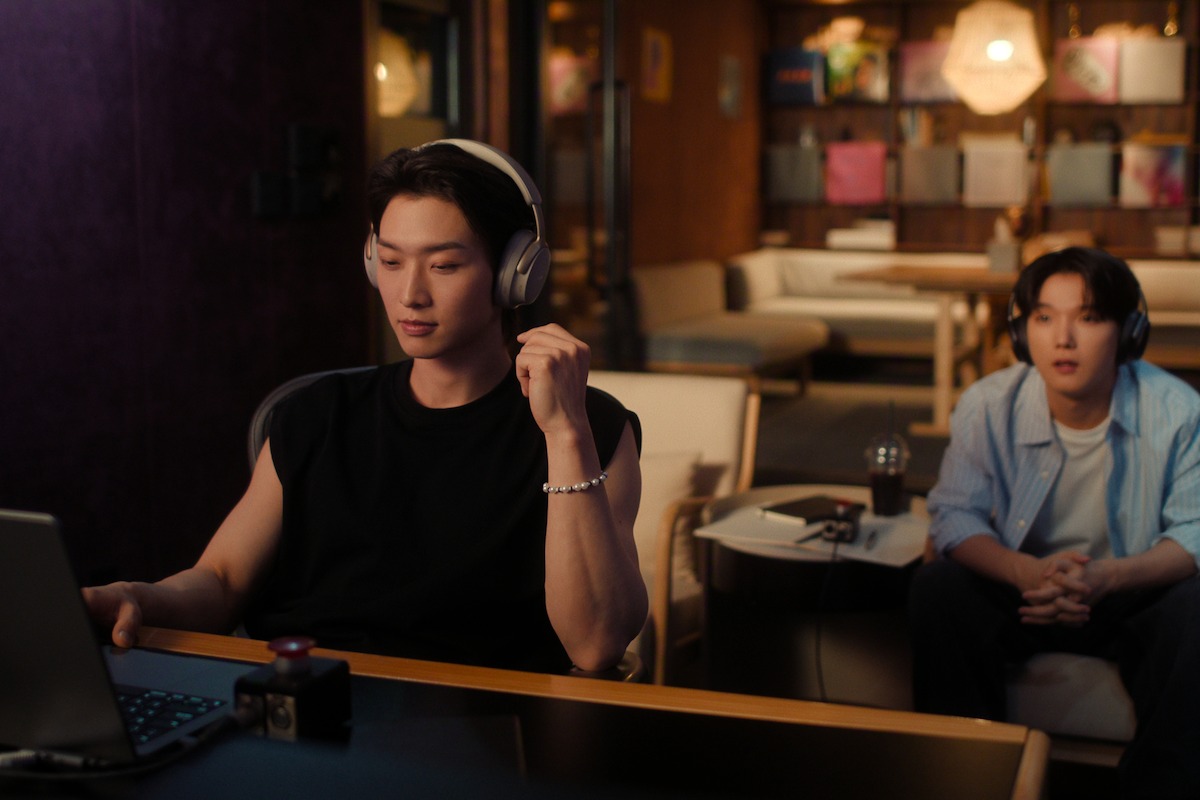 Two people in a cozy, modern room; one in headphones works on a laptop at a desk, the other sits on a sofa with drinks and a notebook on the table, warm lighting and shelves in the background.