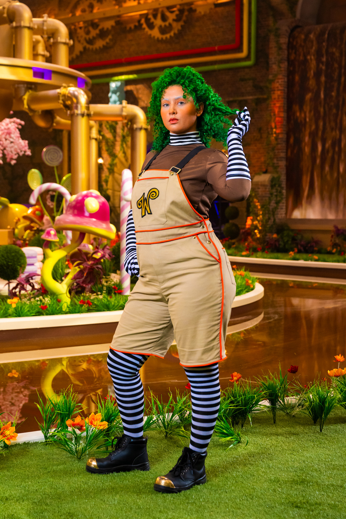 Person dressed as an Oompa Loompa with green hair, striped shirt, and overalls stands confidently in a whimsical, colorful candy factory setting with oversized pipes, mushrooms, and vibrant plants.