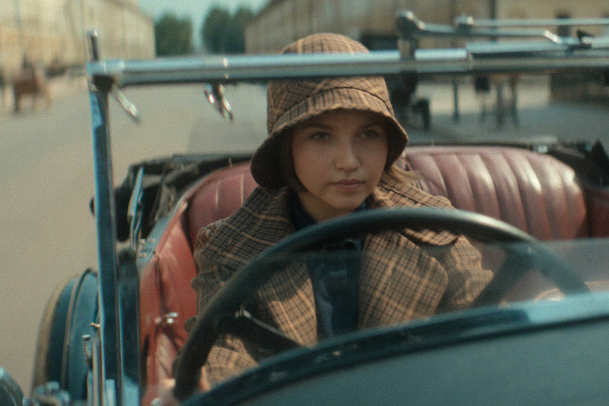 A young woman wearing a plaid hat and coat drives a vintage convertible car with a red leather interior down an empty street lined with buildings, evoking a historical or early 20th-century setting.