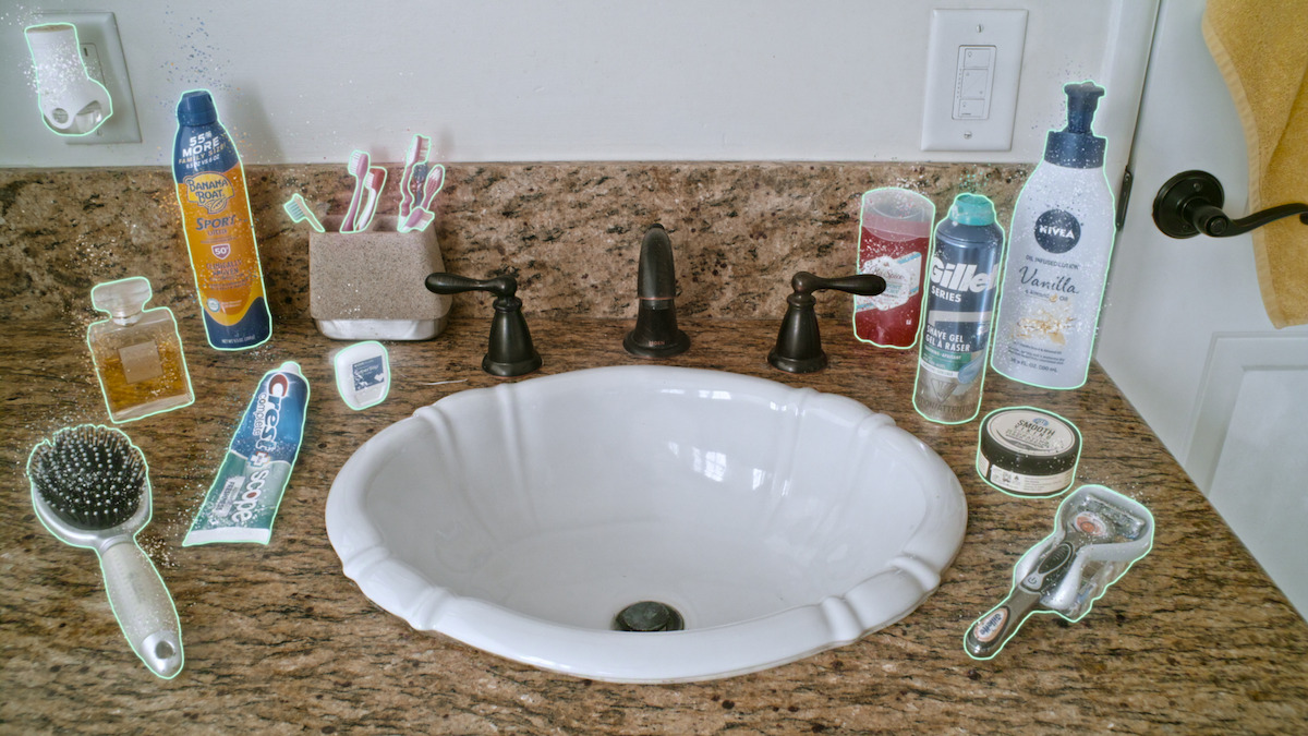 Bathroom sink with brown granite countertop, surrounded by grooming items like razor, brush, toothpaste, toothbrushes, soap, mouthwash, deodorant, shaving foam, lotion, and a towel hanging on the wall.