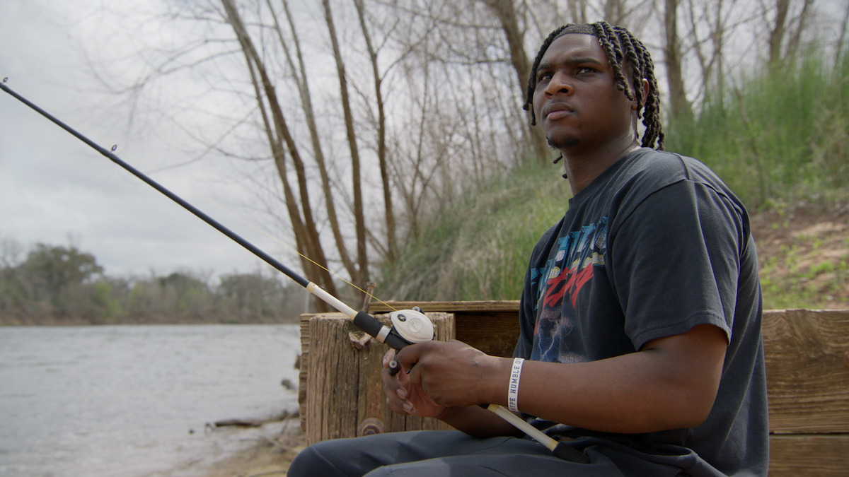 Young man fishing by a riverbank, sitting on a wooden bench surrounded by trees and greenery on a cloudy day, holding a fishing rod and looking thoughtful.