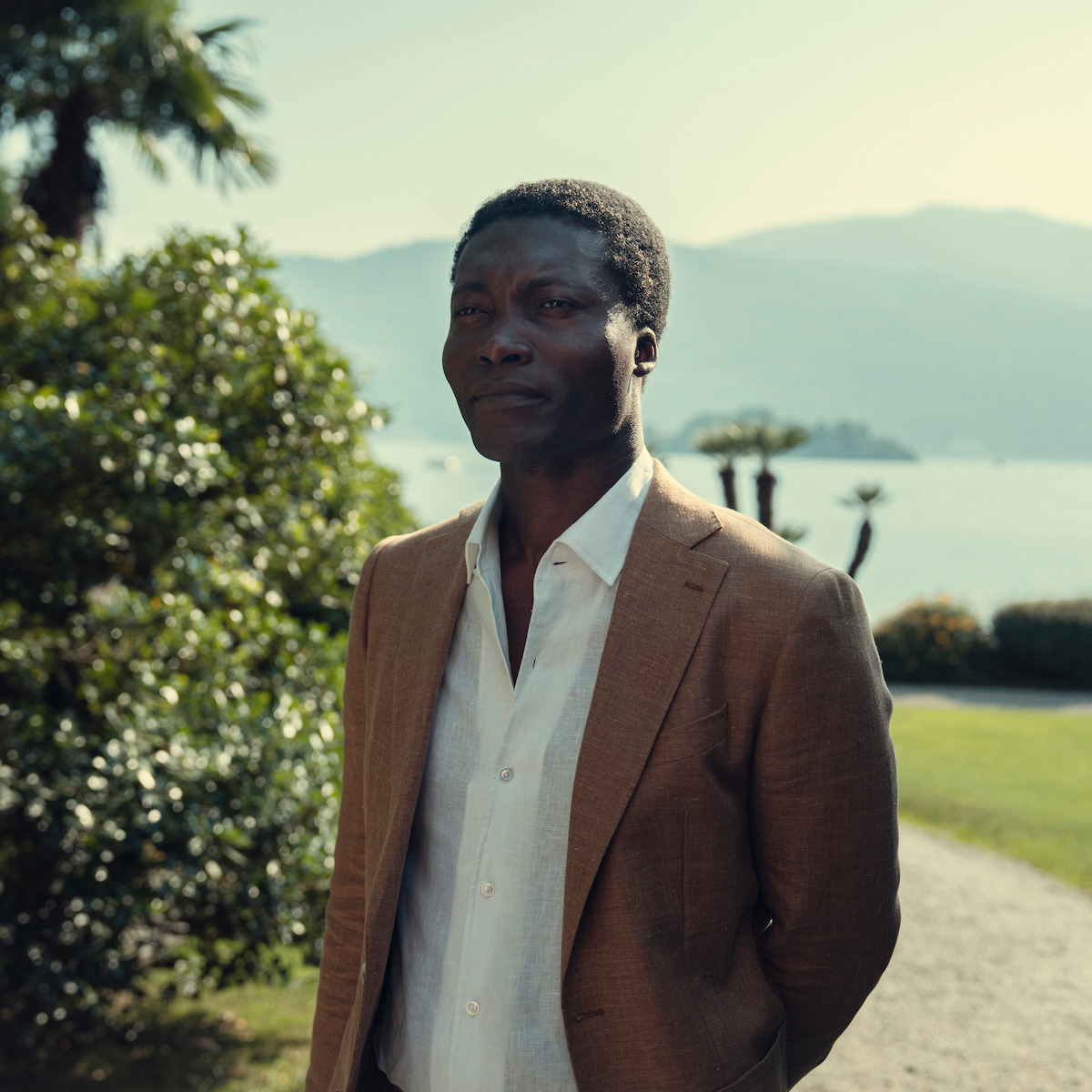 Man in a light brown suit and white shirt standing outdoors on a sunny day with greenery, palm trees, mountain lake, and distant hills in the background.