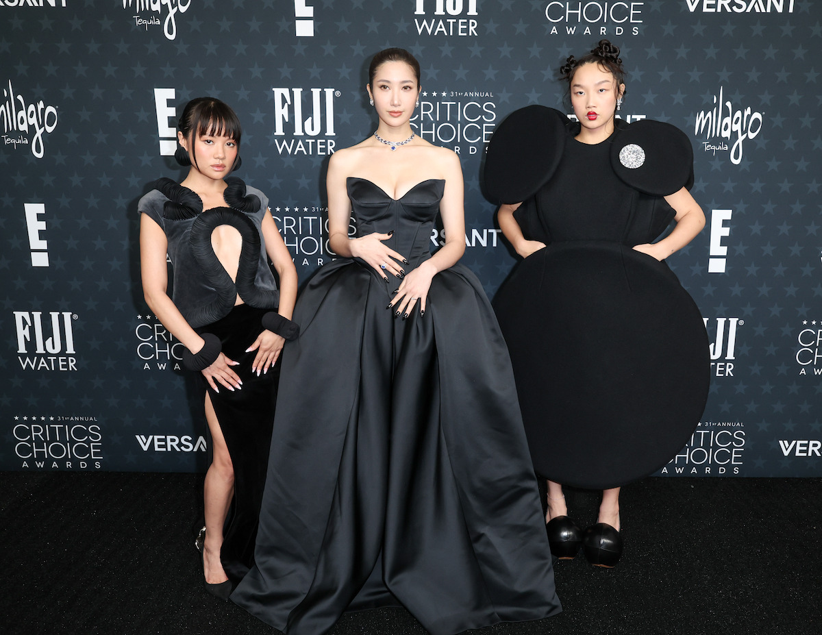 Three women in avant-garde black dresses pose on the red carpet at an awards event, standing before a backdrop with logos like FIJI Water and Critics Choice Awards. The setting is formal and stylish.
