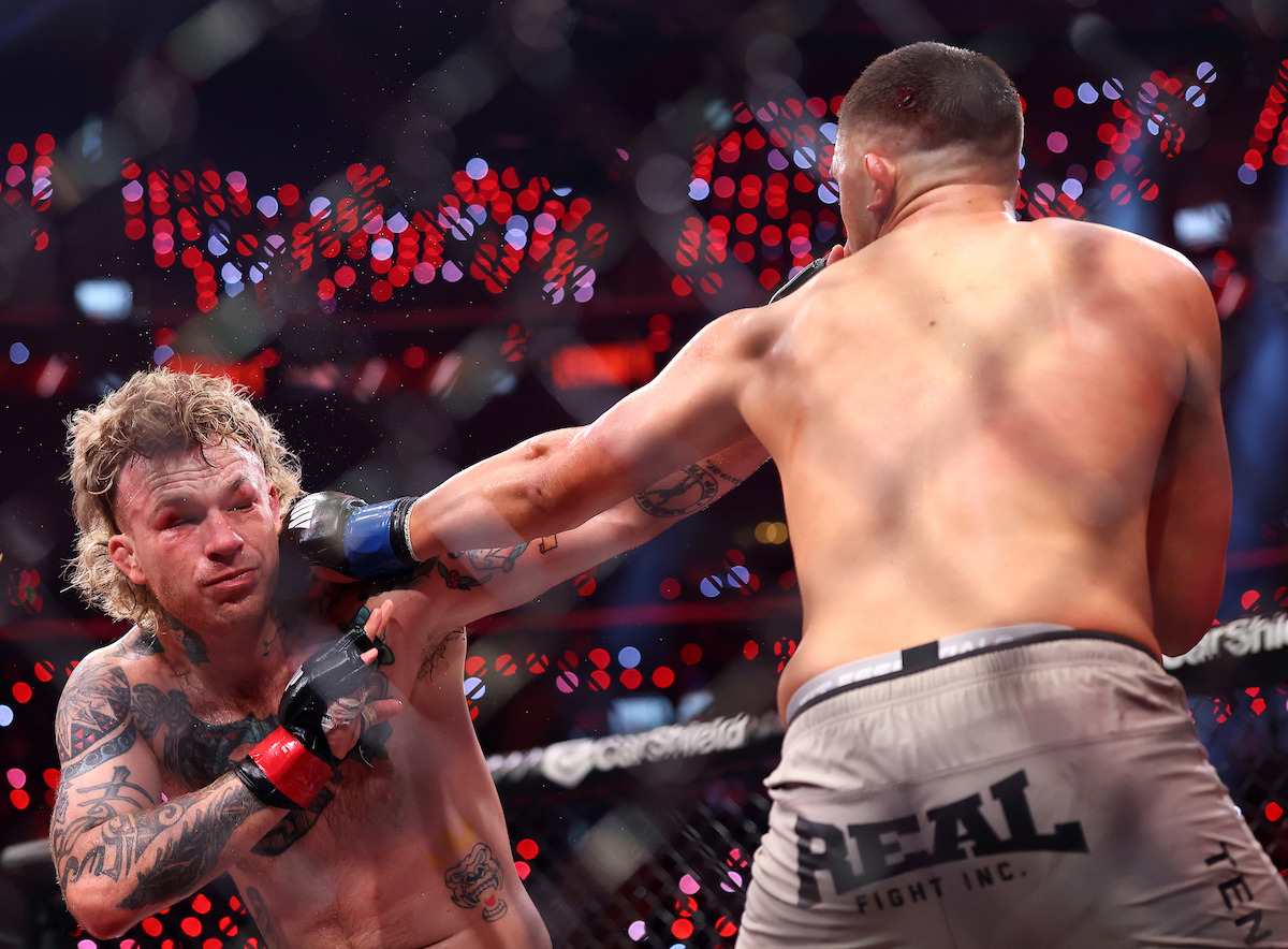Two male MMA fighters engaged in a match inside a cage, one landing a punch to the other's face, under bright arena lights with a crowd in the background.