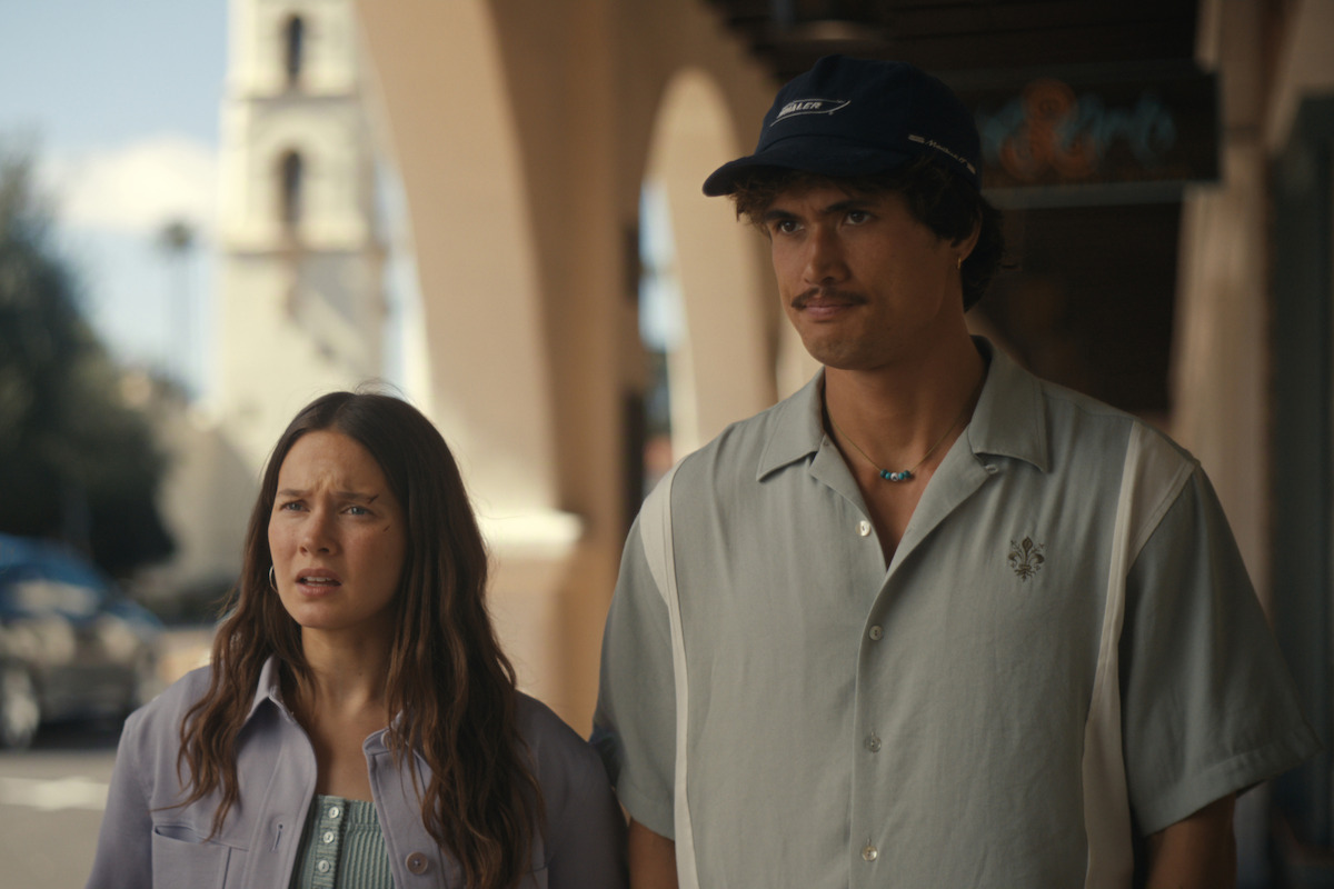 A woman and man stand outdoors on a sunny street with arched walkways and a tower in the background, both appearing concerned or focused.