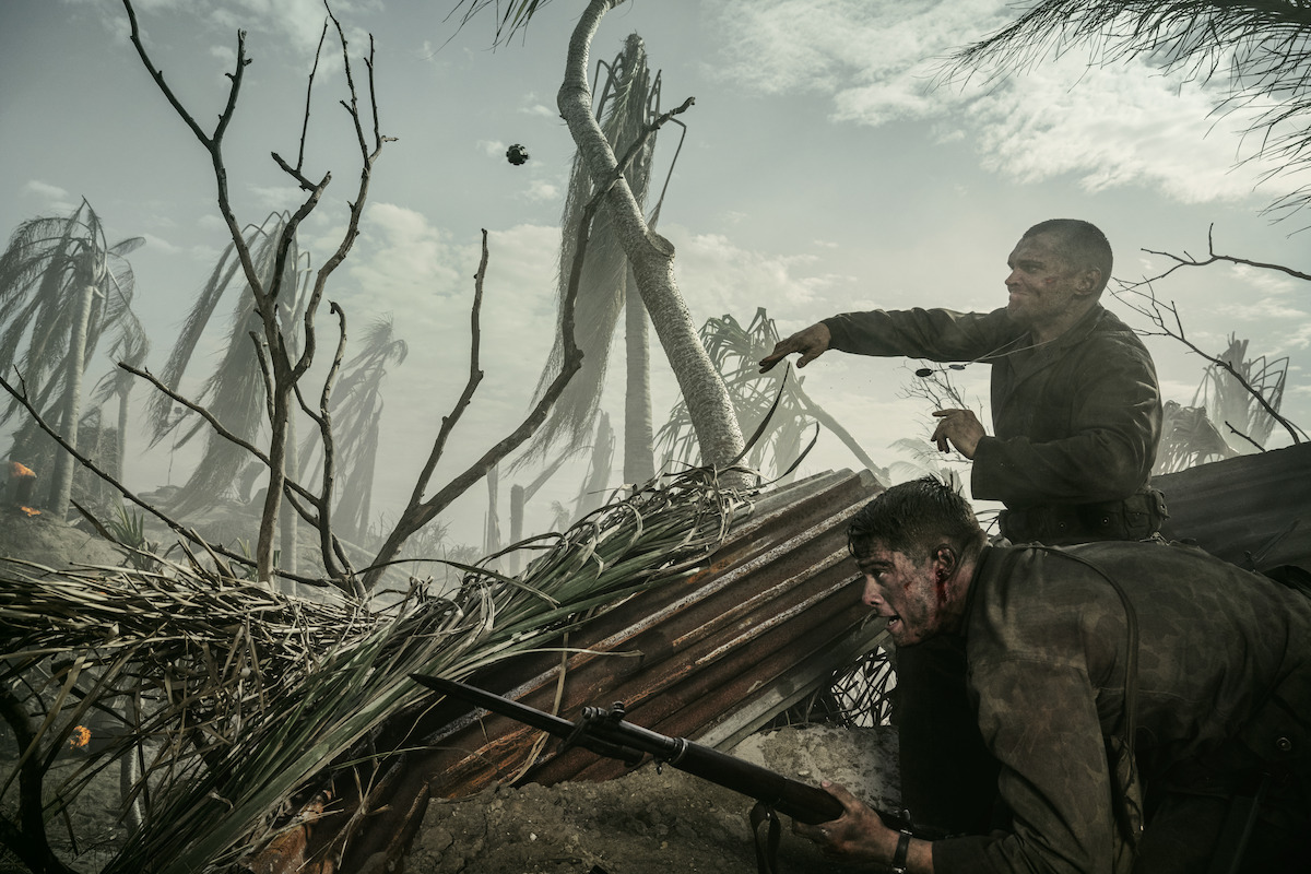 Two soldiers in muddy uniforms take cover behind debris in a war-torn, smoky battlefield with broken trees, holding rifles and looking alert, surrounded by chaos and destruction.