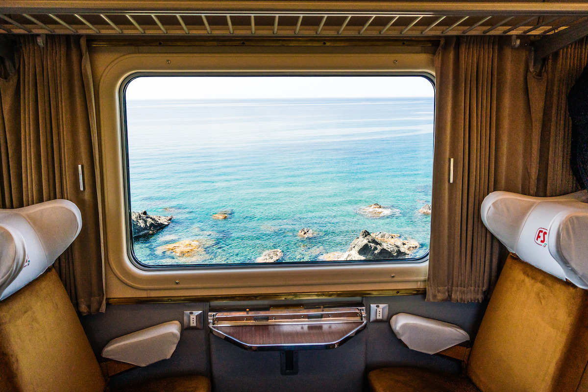 Two empty train seats face a window with a serene view of blue ocean and rocky shore, surrounded by brown curtains. The scene is calm and inviting, suggesting a peaceful journey by the sea.