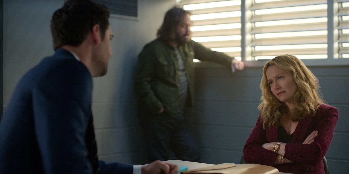 A woman sits at a table with arms crossed, looking serious, while a man in a suit sits across from her and another man stands in the background. The setting appears to be an interrogation room with barred windows.