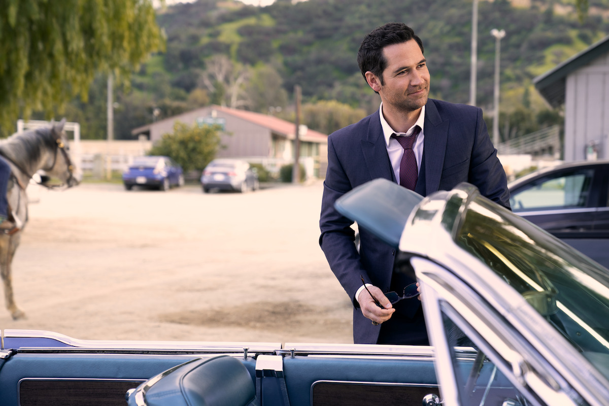 Man in a suit standing beside a classic convertible car in a rural parking lot, with a horse and stable buildings in the background, surrounded by hills and greenery.