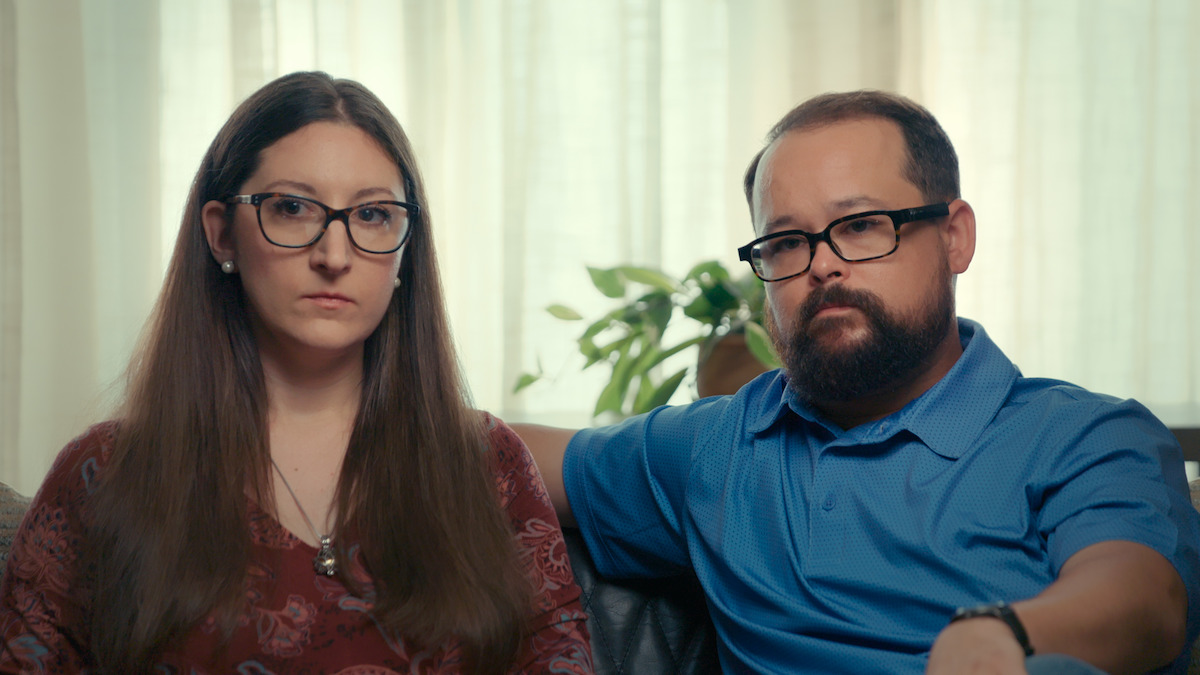 A woman and a man sit closely together indoors on a couch, both wearing glasses and looking serious. The background features soft natural light through curtains and a green potted plant.