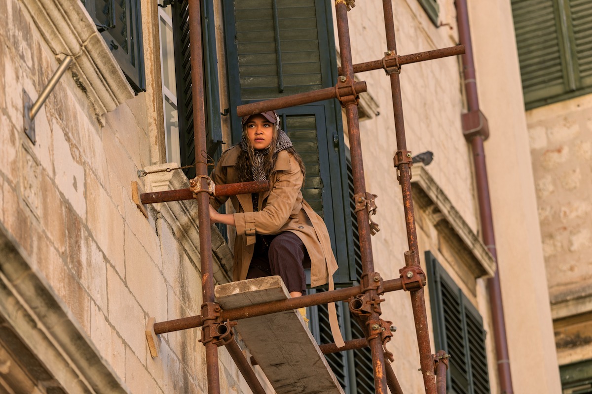 A woman in a brown coat and hat climbs scaffolding attached to the exterior of a stone building with green shutters, appearing cautious and focused in an urban setting.