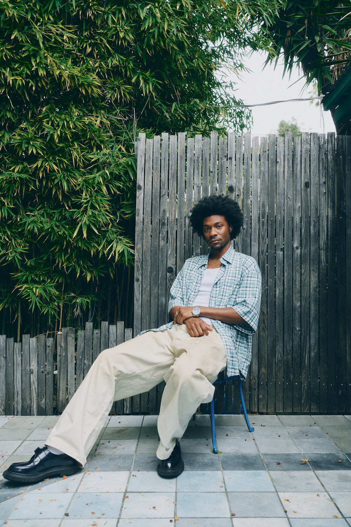 Man with curly hair sits casually on a chair outdoors, wearing a plaid shirt and beige pants, against a wooden fence surrounded by green bamboo plants on tiled flooring.