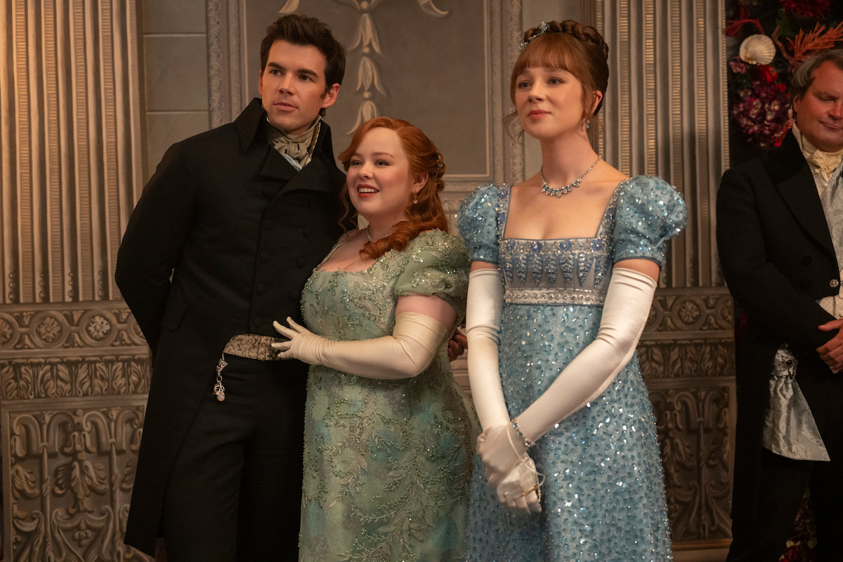 Three people in elegant Regency-era costumes stand together in a lavish, ornately decorated room with stone and floral details, suggesting a formal social gathering or ball.
