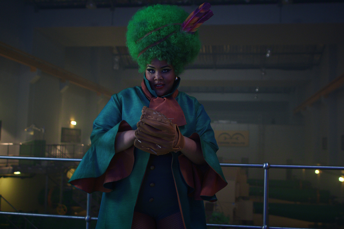 A person with long hair and a tall feathered hat wields a cane or staff menacingly in a dimly lit room, with maps on the wall in the background, creating a mysterious, dramatic atmosphere.