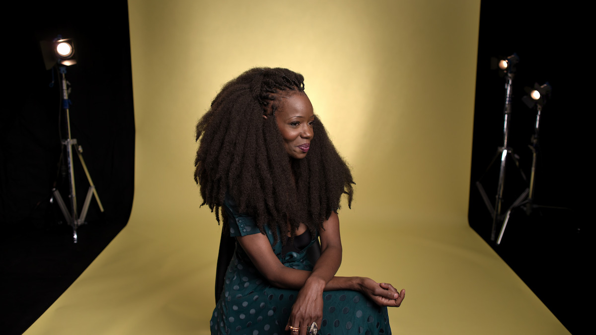 Woman with voluminous natural hair sitting and smiling in a photography studio, dressed in a green dress with polka dots, yellow backdrop and studio lights visible in the background.